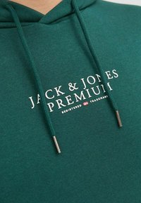 Tyrkysová mikina s kapucňou a sťahovacími šnúrkami, so striebornými kovovými koncovkami, s potlačou loga "JACK & JONES PREMIUM" na hrudníku v bielej farbe. Mäkkučký, textúrovaný materiál.