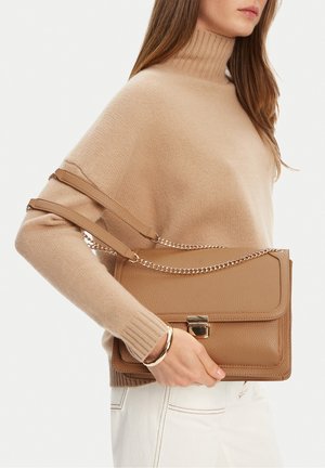 Donna che indossa un maglione beige a collo alto e pantaloni bianchi, con una borsa in pelle marrone chiaro con catena e chiusura dorate, polso adornato da un braccialetto d'oro.
