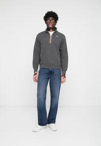 Sweat-shirt en polaire gris avec un zip quart, arborant un col rayé multicolore, associé à un jean usé bleu clair et des baskets blanches.