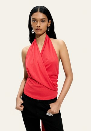 Młoda kobieta z długimi czarnymi włosami, ubrana w czerwony bezrękawnik z dekoltem halter i czarne spodnie, z dodatkami w postaci srebrnych kolczyków i pierścionków.