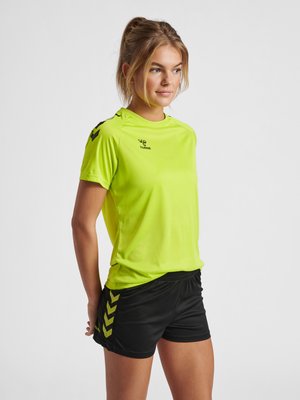 Vrouw in een geel sportshirt en zwarte korte broek staat met haar handen achter haar rug, licht naar rechts kijkend tegen een egale lichte achtergrond.