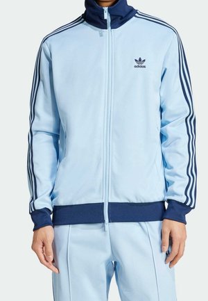 Homme portant une veste de survêtement Adidas bleu clair avec col, poignets et taille bleu foncé, ainsi que trois bandes sur les manches, associée à un pantalon assorti.