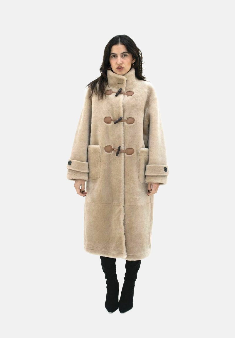 Beige fuskpälsjacka med hög krage, stora framfickor, bruna knappslutningar och breda ärmar. Buren över svarta knästövlar.