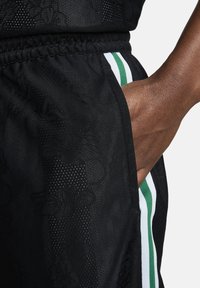 Bermudas deportivas negras con una textura de malla, que presentan un patrón sutil. Bolsillos laterales con bordes en rayas verdes y blancas. Cintura elástica.