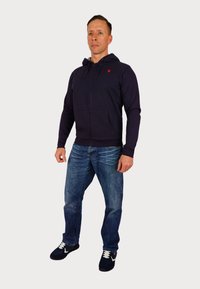 Námornícky zipsovaný mikina s prednými vreckami, nosená s modrými džínsami a čiernymi športovými topánkami. Mikina má nastaviteľné šnúrky a akcent loga.