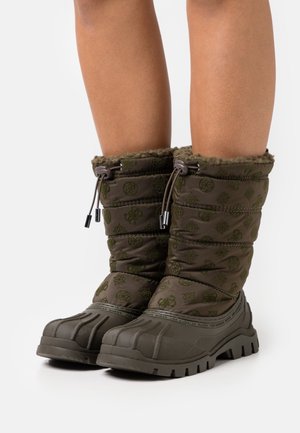 Persona que lleva botas de invierno acolchadas de color verde oscuro con tela estampada, cierres con cordón y suelas de goma resistentes sobre un fondo blanco.