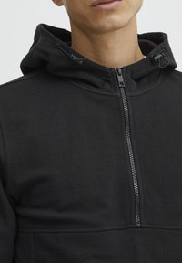 Svart hettegenser med strukturert stoff, glidelås og justerbare snorer i hetta. Har høy hals og minimalistisk design.