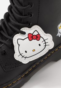 Černá kožená bota s žlutým prošíváním, která má bílou nášivku s obličejem Hello Kitty a červenou mašlí připevněnou poblíž šňůrek.