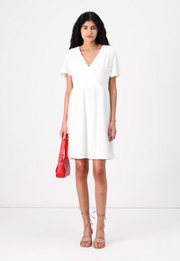 White short-sleeve wrap dress with textured fabric, above knee length, paired with a red handbag and strappy nude sandals.