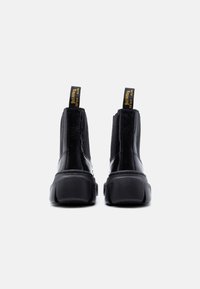 Γυαλιστερά μαύρα μποτάκια αστραγάλου με χοντρές σόλες και κίτρινες ταινίες έλξης που αναγράφουν «WITH SOLES BOUNCING», εμφανιζόμενα από πίσω σε λευκό φόντο.