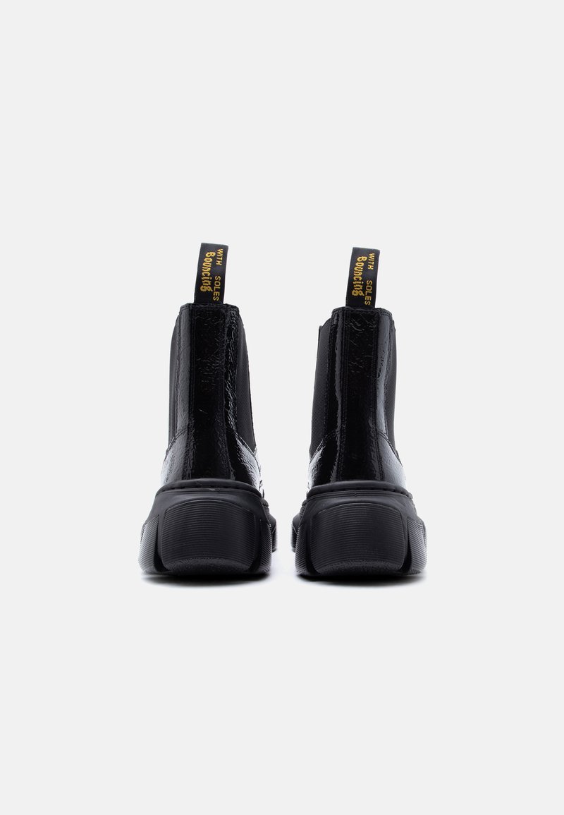 Glanzende zwarte enkellaarsjes met dikke zolen en gele aantreklussen met de tekst "WITH SOLES BOUNCING", vanaf de achterkant getoond op een witte achtergrond.