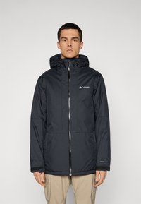Man wearing black Columbia jacket with hood and front zipper, standing against plain light background.