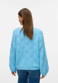 Hellblauer Strickpullover mit Rautenmuster, lockere Passform, übergroße Ärmel und gerippter Ausschnitt. Getragen mit blauen Jeans, die minimalen Texturen zeigen.