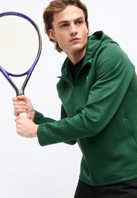 Νέος άντρας με πράσινο φούτερ με φερμουάρ που κρατά ρακέτα τένις, έτοιμος να χτυπήσει, με φόντο απλό λευκό.