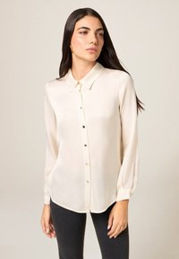 Beige blouse met lange mouwen, een knopenrij aan de voorkant, een puntige kraag en een subtiele glans. Voorzien van kleine, afgeronde knopen en een afgeronde zoom.