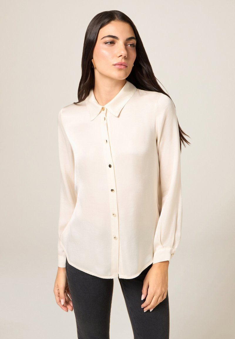 Beige blouse met lange mouwen, een knopenrij aan de voorkant, een puntige kraag en een subtiele glans. Voorzien van kleine, afgeronde knopen en een afgeronde zoom.
