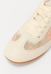 Baskets beiges avec une tige en cuir lisse, des panneaux en mesh ornés de broderies florales roses, et des lacets plats en satin. Bout arrondi et semelle en caoutchouc.