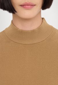 Gros plan d'une personne aux cheveux courts et foncés portant un pull à col montant côtelé de couleur camel.