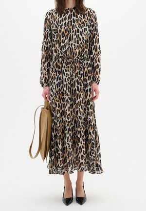 Nainen pukeutuneena pitkähihainen, nilkkoihin ulottuva leopardikuvioinen mekko, kädessään vaaleanruskea käsilaukku ja jalassaan mustat teräväkärkiset korkokengät, seisoo valkoista taustaa vasten.