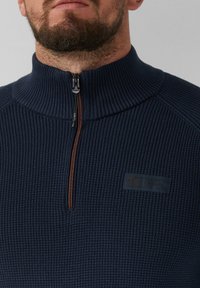Marineblauer, strukturierter Pullover mit hohem Kragen, halbseitigem Reißverschluss und einem kleinen Logo-Patch auf der Brust. Leichtes Strickmaterial.