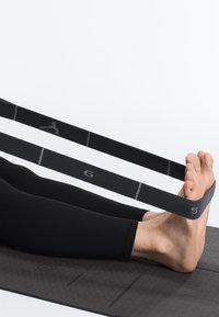 Personne utilisant une sangle noire d’étirement numérotée 5 et 6 autour du pied nu, assise sur un tapis d’exercice noir texturé avec les jambes tendues.