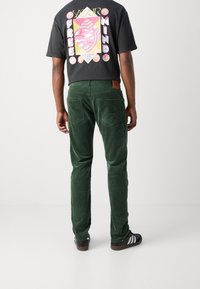 Person som bär mörkgröna manchestertights, svart grafisk t-shirt med texten "Your Free Mind" och svarta sneakers med vita ränder, stående med ryggen mot kameran.