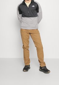 Pullover nero e grigio con mezza zip, realizzato in tessuto testurizzato. Pantaloni cargo color beige con tasche laterali. Scarpe da trekking nere con lacci.