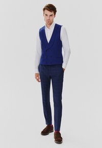 Niebieska kamizelka podwójnie zapięta na biały shirt, w połączeniu z dopasowanymi granatowymi spodniami w kratkę i brązowymi butami. Prosty, dopasowany krój.