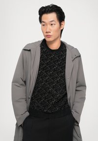 Manteau gris sur un pull noir à motif, présentant un tricot texturé. Le pull a un col rond et est associé à un pantalon sombre.