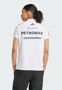 Hvid polo-shirt med krave, med sorte sponsorlogoer: PETRONAS og Crowdstrike, samt sorte accenter på skuldrene.