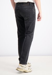 Mustad denim püksid kitsas lõikes, tagakotid ja nähtav brändi silt vöö peal. Seljas hallid ja valged tossud.