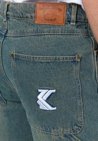 Denimplagg i mediumblått med en läderlapp vid midjan och en broderad logotyp på bakfickan. Klassisk rak ben-design.