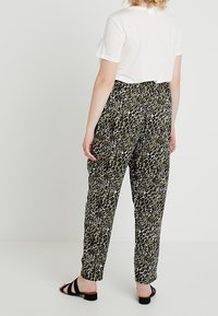 Luipaardprint broek in groen, zwart en wit; losse pasvorm met een elastische tailleband; model draagt een wit T-shirt met korte mouwen en zwarte sandalen.
