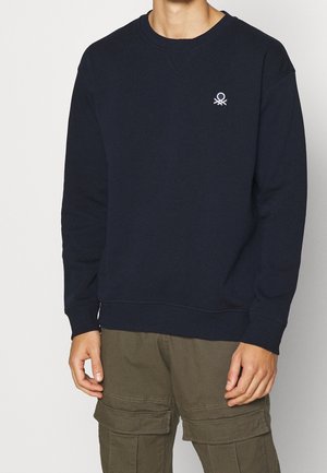 Persona vistiendo una sudadera azul marino con un pequeño logo blanco en el pecho y pantalones cargo color verde oliva con múltiples bolsillos.