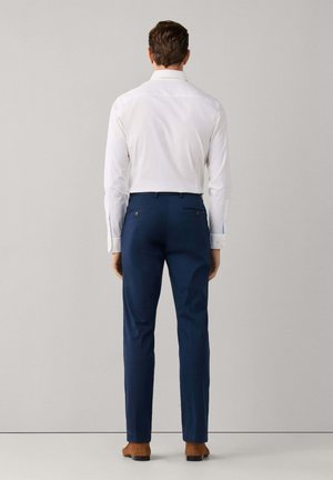 Homme portant une chemise blanche, un pantalon bleu marine et des chaussures marron, debout face à un mur gris clair uni.