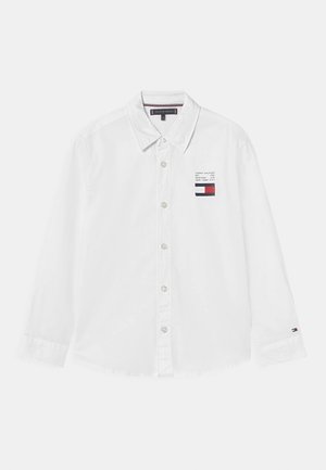 Camisa blanca de manga larga con cuello abotonado, que presenta un pequeño parche de logo en colores rojo, blanco y azul en el lado izquierdo del pecho.