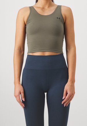 Kvinde iført en tætsiddende olivengrøn ærmeløs crop top og mørkeblå leggings med høj talje står mod en ensfarvet baggrund.