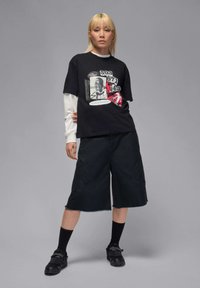 Camiseta gráfica negra con mangas largas blancas, combinada con pantalones cortos oscuros de dobladillo irregular, calcetas negras y zapatos negros. Diseño minimalista.