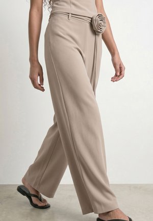 Pantalon beige à jambes larges avec une ceinture en tissu nouée en forme de rose à la taille, porté avec des sandales plates noires.