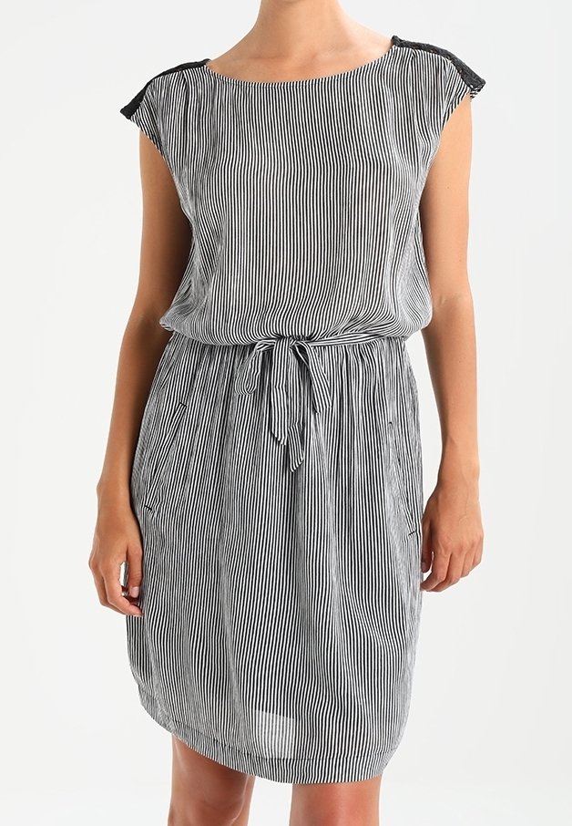 Femme portant une robe sans manches rayée verticalement noir et blanc, longueur genou, avec une ceinture nouée à la taille et une taille froncée.