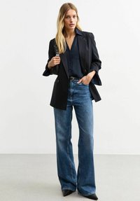 Blazer nero a righe sottili sopra una blouse blu navy, abbinato a jeans a gamba larga blu chiaro e tacchi a punta neri. Tessuto liscio e forma sartoriale.