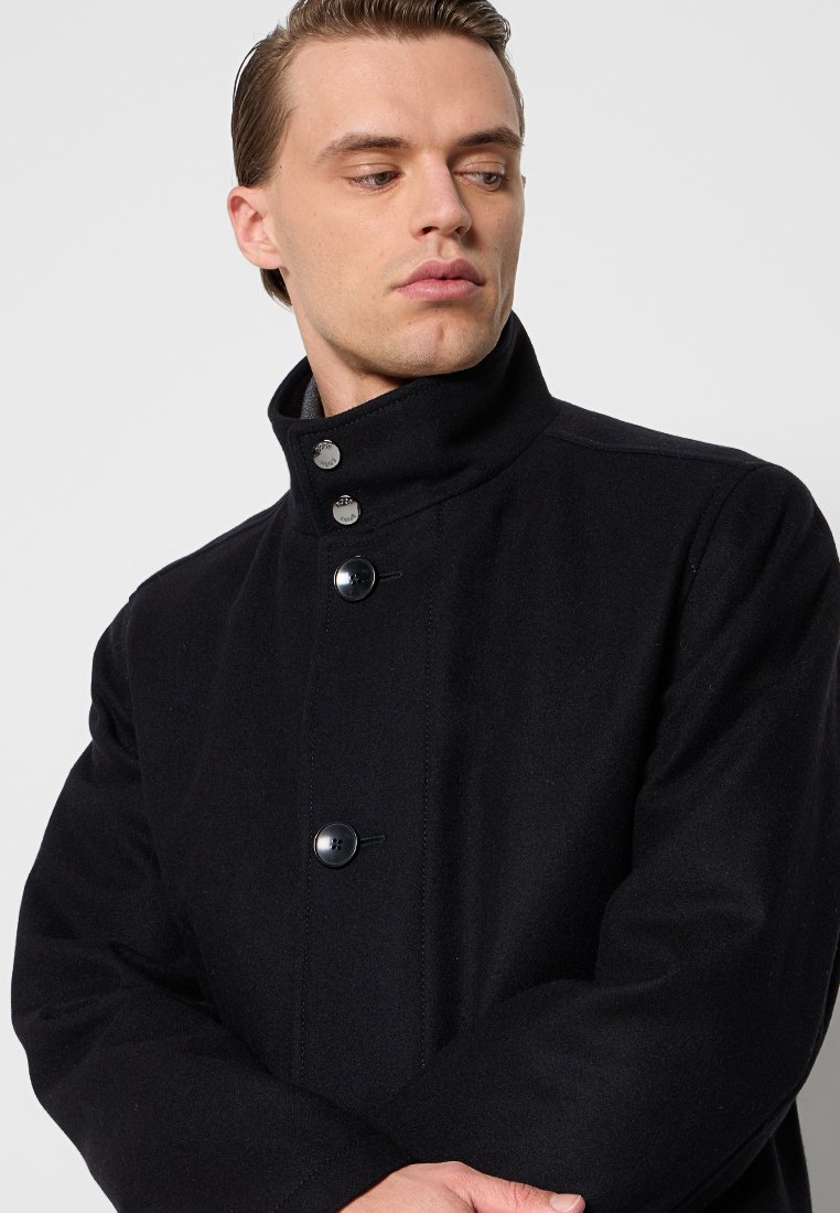 Manteau en laine noir avec un col haut, comportant quatre grands boutons et une texture lisse, taillé avec un design ajusté.