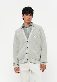 Light gray knit cardigan with a V-neck, textured fabric, black buttons, and two front pockets. Worn over a blue striped shirt.