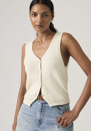 Mujer con chaleco sin mangas de punto color crema abotonado y jeans de cintura alta azul claro con un cinturón marrón, posando contra un fondo liso.