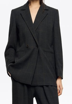 Blazer croisé à carreaux gris foncé et pantalon large assorti porté par une personne avec la main dans la poche.