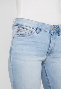 Jeans de mezclilla azul claro con un corte Slim, un bolsillo frontal con cremallera y una cintura clásica con cierre de botón. Textura suave, desvanecimiento mínimo.