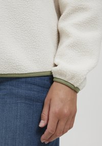 Fleecová mikina v off-white farbe s textúrovaným povrchom, zdobená zeleným lemovaním na rukávoch a spodnom okraji, v kombinácii s tmavými džínsy.
