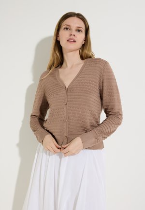 Vrouw die een bruine, getextureerde cardigan met ribbels dichtknoopt, een witte rok draagt en voor een effen lichte achtergrond staat.