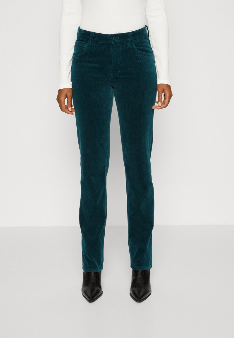 Personne portant un pantalon en velours côtelé vert foncé, un haut blanc à manches longues et des bottines noires à bout pointu, debout sur un fond uni.