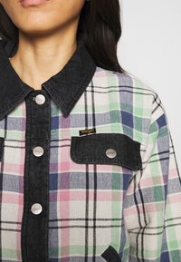 Femme portant une veste-chemise à carreaux avec des carreaux pastels verts, roses et bleus, col en denim noir, patte de boutonnage et rabat de poche avec logo Wrangler.